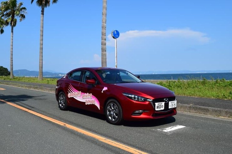 海沿いの道を走る蒲郡自動車学校の教習車